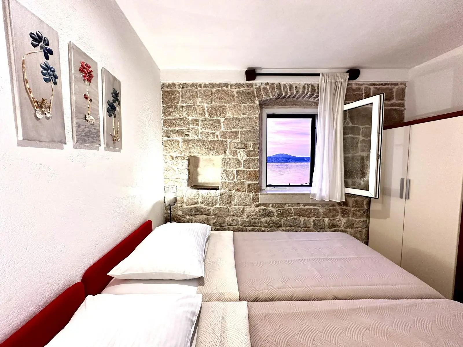 Modern bedroom with a large bed, stone walls, chic decor, and a window offering a view of the sea at dusk.