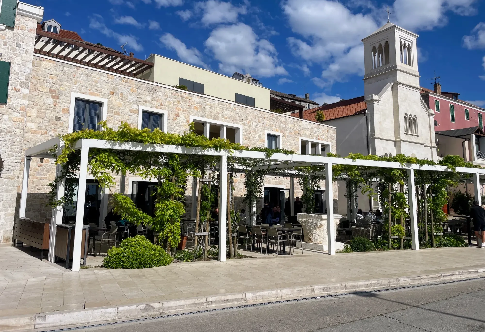 A picturesque outdoor cafe with a pergola and greenery, nestled beside a historic stone building and a church tower under a blue sky.