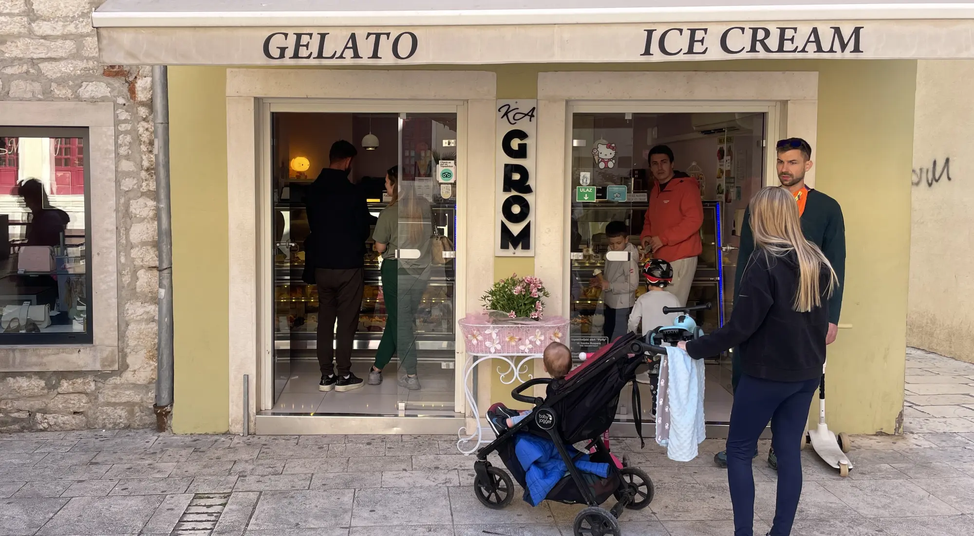 People visiting a 'Gelato Ice Cream' shop with stone façade, as a woman with a stroller waits outside