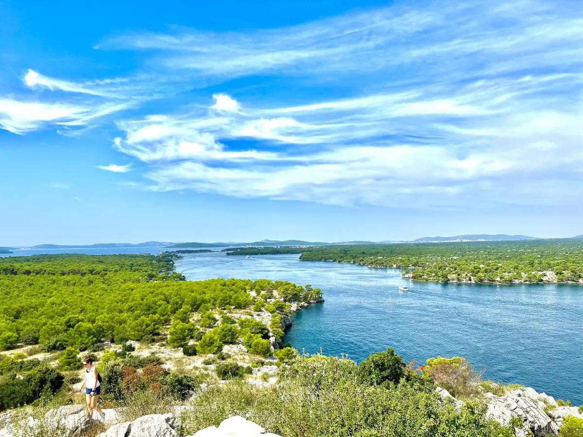 Panoramic view from a high vantage point overlooking the channel between the dense, green forested islands and the mainland, under a vivid blue sky with wispy clouds. A person is visible on the rocky foreground, taking in the scenery.