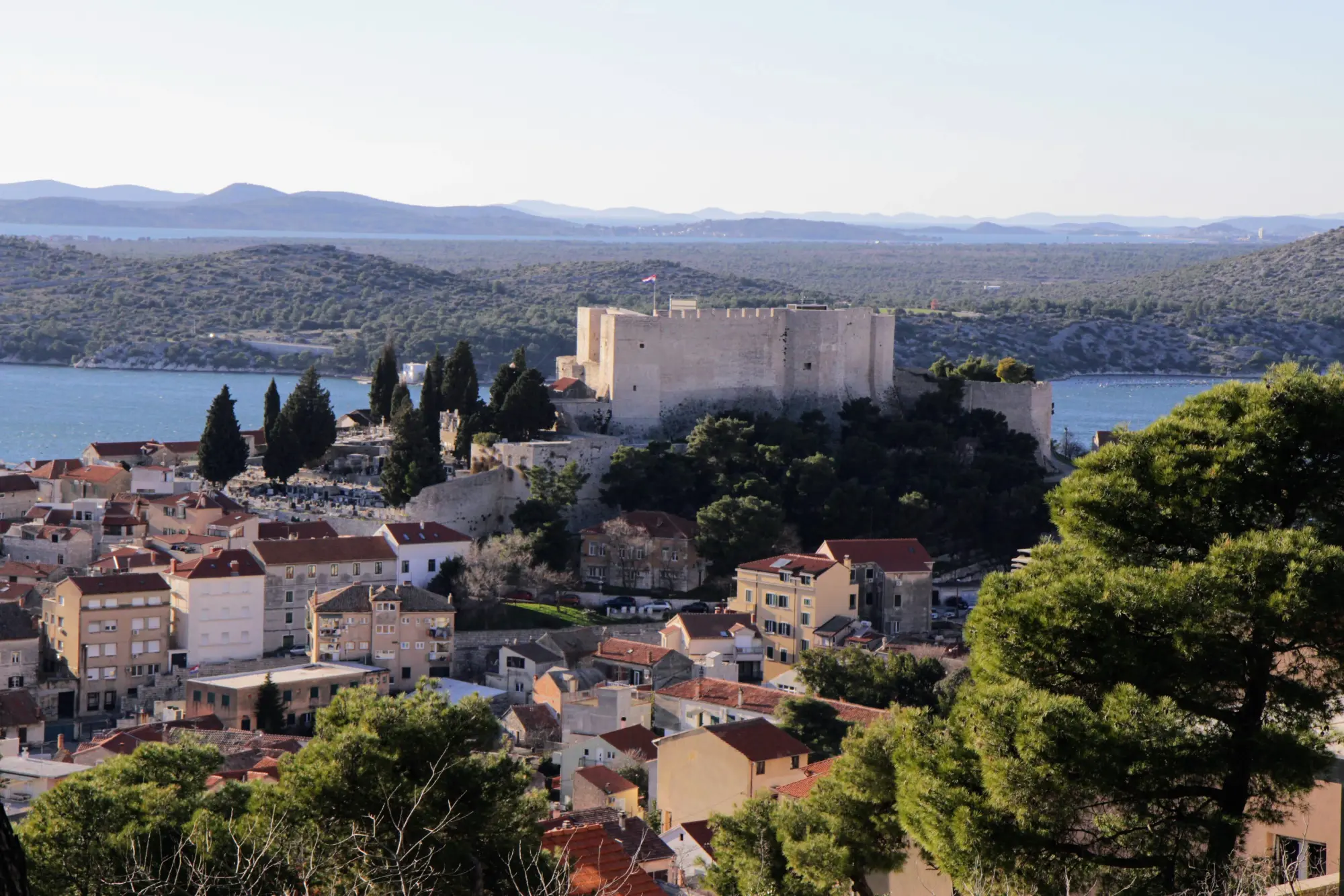 Aerial view of the historic town of Šibenik during the golden hour, showcasing the dense clustering of red-roofed houses, the prominent cathedral, and the marina filled with boats along the calm Adriatic coast.