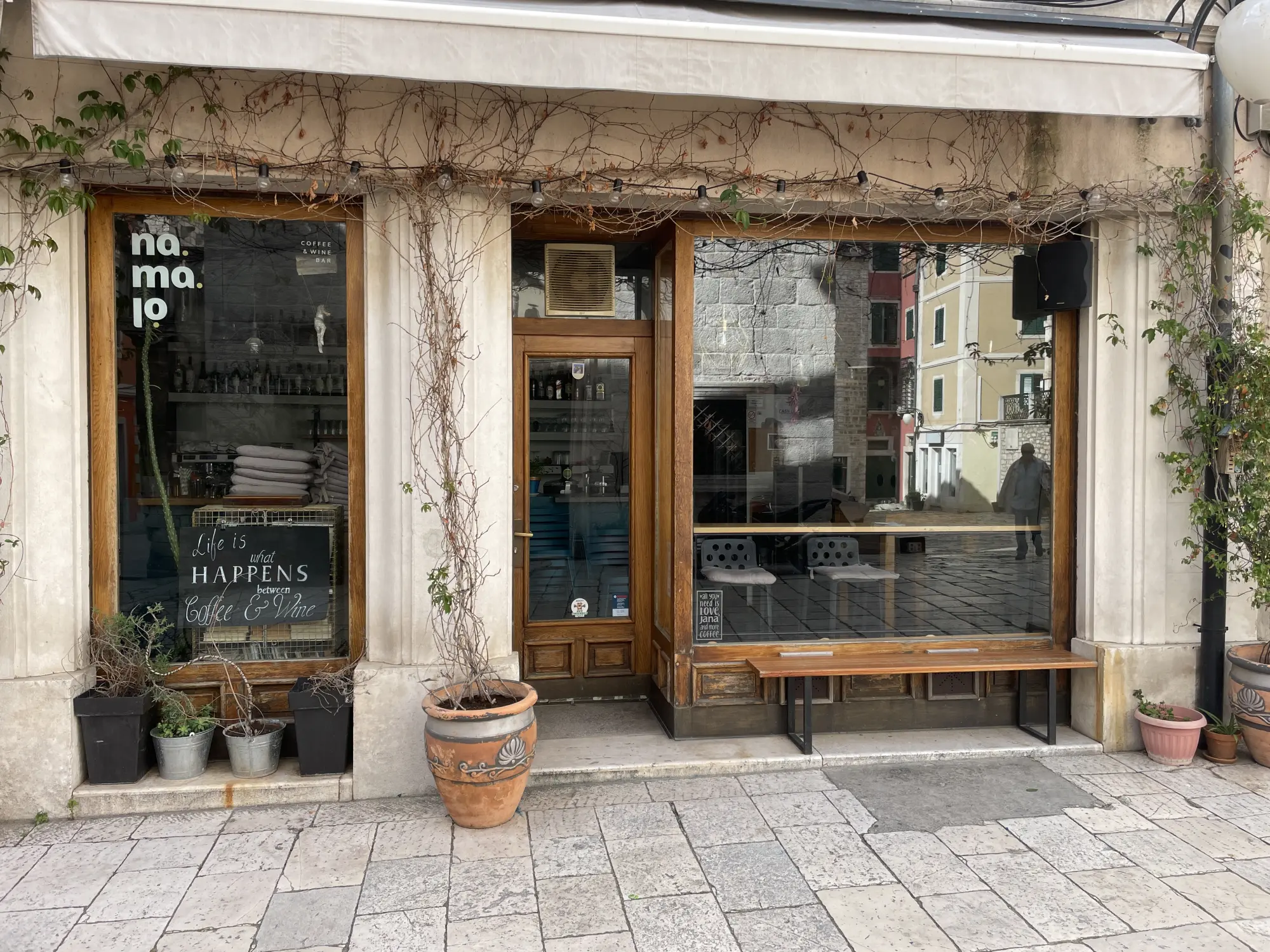 Cozy street-level cafe facade with vine overhangs and a chalkboard sign, inviting passersby for coffee and wine.