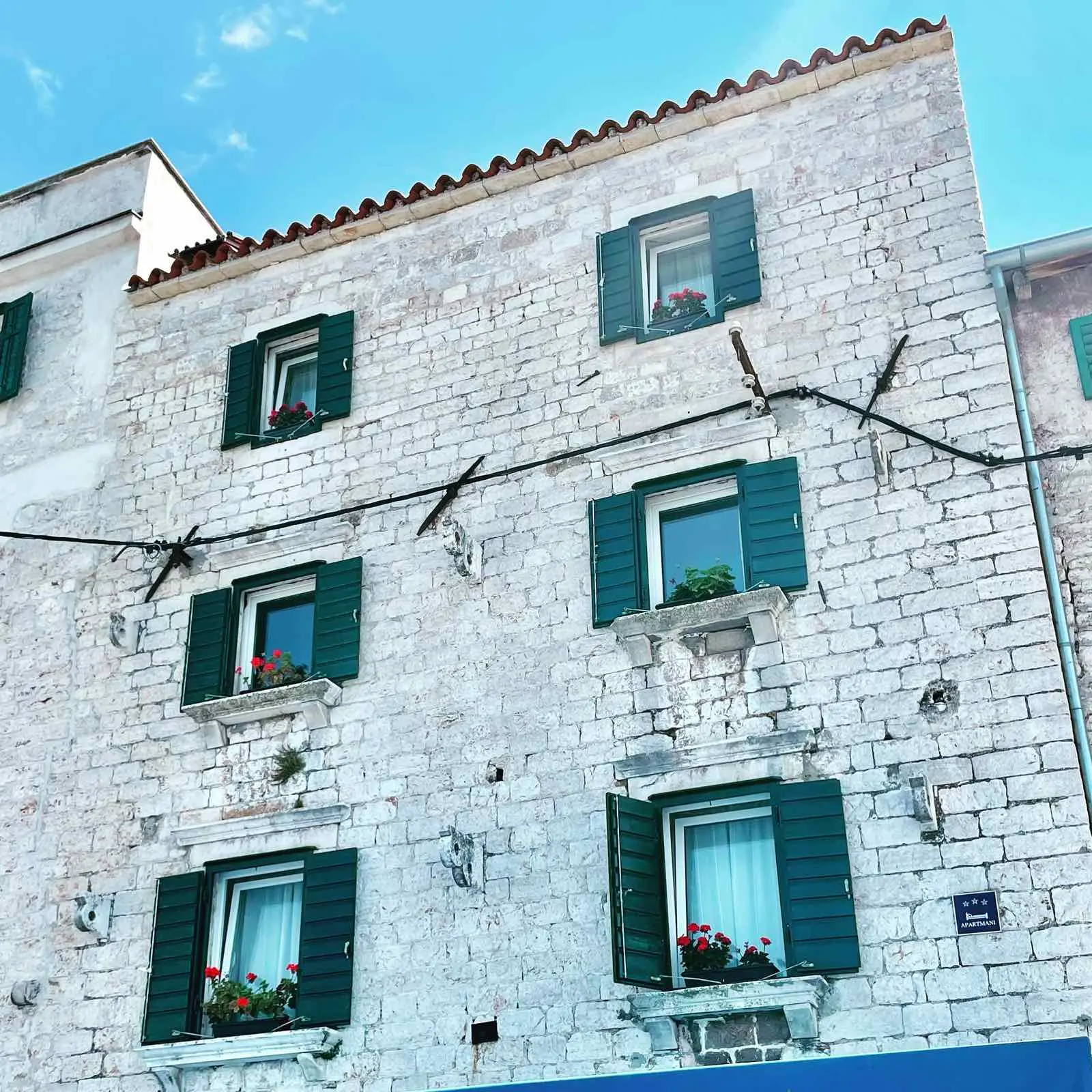 Historic stone building with green shutters and blooming flowers on the windowsills against a clear blue sky